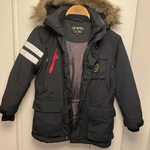 Svart parkajacka från Gneis med päls 122 - Parkajacka från Gneis i svart med fuskpäls på huvan, reflexdetaljer på ärmen och röd dragkedja på bröstfickan. Jackan har flera fickor, justerbar midja och långärmad design. Perfekt för kalla dagar och outdoor-aktiviteter. Reflexen där bak saknas tyvärr men annars super fin