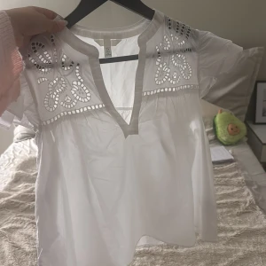 Vit blus med broderade detaljer H&M - Superfin vit blus från H&M med broderade detaljer och hålmönster upptill. Blusen har v-ringning, volangiga korta ärmar och är gjord i ett lätt bomullstyg. Perfekt för en fräsch och luftig look under varmare dagar.