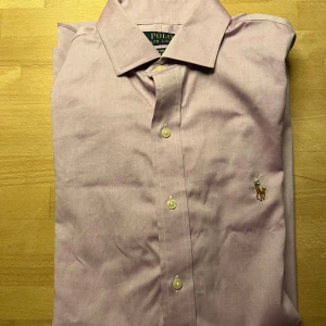 Ljuslila skjorta från Polo Ralph Lauren - Säljer en ljuslila skjorta från Polo Ralph Lauren med klassisk krage och broderad logga på bröstet. Den har knappar framtill och är i bomull. Custom fit-modell som sitter snyggt och stilrent.    Strl 15/38