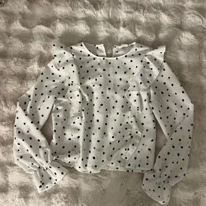Vit prickig blus med volang KappAhl - Supersöt vit blus från KappAhl med svarta prickar och volangdetaljer längs axlarna. Långärmad modell med lätt puff vid ärmslut och rund halsringning. Perfekt för dig som gillar en lekfull och trendig stil.