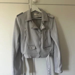 Ljusgrå bikerjacka - Snygg ljusgrå bikerjacka med klassisk krage, axelklaffar och flera dragkedjor framtill. Jackan har bälte i midjan och är tillverkad i mjukt material som ger en cool look. Perfekt för dig som vill ha en stilren och trendig jacka.