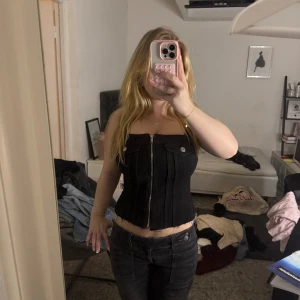 Svart jeanskorsett med dragkedja - Säljer en svart korsettopp i jeansmaterial med markerade sömmar och rå kant nertill från Zara. Toppen är axelbandslös och har en snygg silverfärgad dragkedja framtill samt dekorativa fickdetaljer med knapp vid bysten. Perfekt för att lyfta vilken outfit som helst.