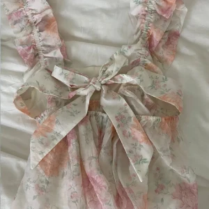 Blommig topp - Supersöt topp från H&M Divided i ljusa toner med blommigt mönster i rosa och persika. Toppen har volangprydda axelband, rosett som man knyter i ryggen och är tillverkad i ett tunt, luftig material