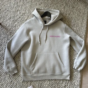 Ljusgrå hoodie från Mads Nørgaard - Ljusgrå hoodie från Mads Nørgaard med rosa logga på bröstet. Klassisk modell med huva, snörning och stor känguruficka framtill. Mjuk insida och ribbade muddar vid ärmslut och nederkant. Perfekt för en chill och stilren look.
