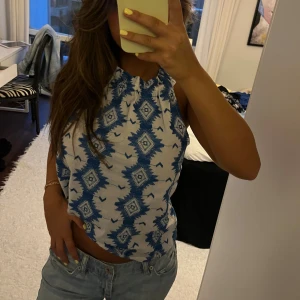 Vit och blå mönstrad linnetopp - Säljer en snygg ärmlös linnetopp i vitt med blått grafiskt mönster. Toppen har halterneck med rynkad halsringning och är lätt croppad. 