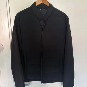 Svart pufferjacka från Zara - Svart pufferjacka från Zara med stilren design och diskret krage. Jackan har raka linjer, quiltad struktur och hel dragkedja framtill. Långärmad och passar perfekt för kyliga dagar. Enkel och snygg look som funkar till många outfits.
