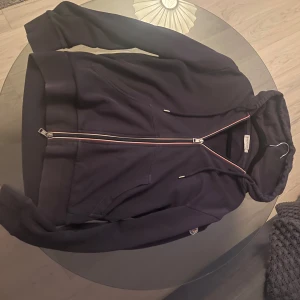Moncler tröja  - Svart hoodie från Moncler med hel dragkedja och huva. Snygga detaljer med röd och vit rand längs dragkedjan. Tillverkad i mjuk bomullsmix och har långa ärmar samt fickor framtill. Perfekt för en chill och stilren look.
