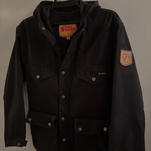 Svart Fjällräven jacka - Passar S/M. Svart parkajacka från Fjällräven i G-1000 material, med huva och flera fickor med tryckknappar. Klassisk Fjällräven-logga på ärmen och snygga detaljer. Perfekt för dig som gillar outdoor och vill ha en stilren jacka med funktion.