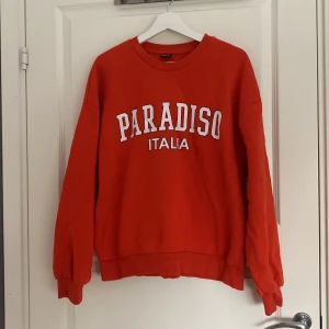 Orange sweatshirt Paradiso Gina Tricot - Orange (ser lite röd ut)  sweatshirt från Gina Tricot i storlek L med vit text 'PARADISO ITALIA' framtill. Tröjan har rund hals, långa ärmar och en loose passform. Perfekt för chill dagar och enkel att styla med jeans eller mjukisbyxor.