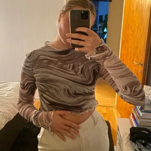 Brun mesh topp med swirlmönster - Säljer en transparent mesh topp från NLY One med swirlmönster i bruna och beige toner. Toppen är croppad, har lång ärm och vågiga kanter vid ärmslut och nederkant.