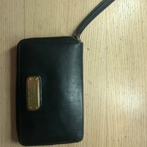 Svart plånbok Marc by Marc Jacobs - Snygg svart plånbok från Marc by Marc Jacobs i äkta skinn med guldfärgad metallbricka framtill. Plånboken har dragkedja runtom, flera kortfack, myntfack med dragkedja och transparent fönster för ID eller busskort. Perfekt att ha med sig överallt.