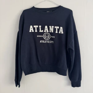 Mörkblå sweatshirt från ONLY med tryck - Mörkblå sweatshirt från ONLY med vit text 'ATLANTA' och collegeinspirerat tryck framtill. Tröjan har rund hals, långa ärmar och ribbade muddar vid ärmslut och nederkant. 