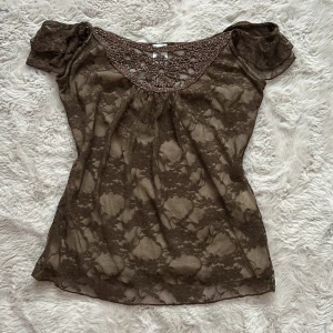 Brun spetsblus med virkad detalj - Säljer en brun, transparent spetsblus med offshoulder-ärmar och virkad detalj vid urringningen. Blusen har blommigt spetsmönster och små volanger på axlarna. Perfekt för dig som gillar romantiska och unika plagg med mycket detaljer. ❤️