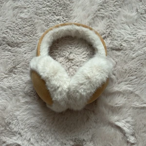 Beige öronmuffar med fuskpäls - Mjuka och varma öronmuffar i beige mockaimitation med fluffig vit fuskpäls runtom. Perfekta för kalla vinterdagar och ger en mysig känsla samtidigt som de håller öronen varma. Snygg och enkel design som passar till det mesta.