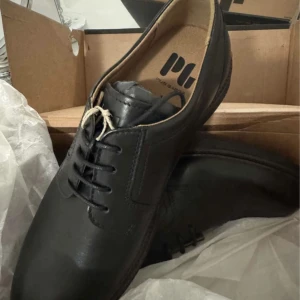 Svarta skinnloafers från Plus Labor - Stilrena svarta loafers från Plus Labor i äkta skinn med klassisk snörning och rund tå. Snygg kontrastsöm och robust platt sula. Perfekta för dig som vill ha en clean och tidlös look till vardags eller mer dressade tillfällen.