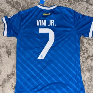Real Madrid Vini Jr. blå matchtröja Adidas - Blå Real Madrid fotbollströja från Adidas med Vini Jr. och nummer 7 på ryggen. Tröjan har vita detaljer, randiga ärmslut och axelband, broderat klubbmärke och Adidas-logga samt Emirates Fly Better-tryck. Tillverkad i lätt polyester med subtila diagonala ränder.
