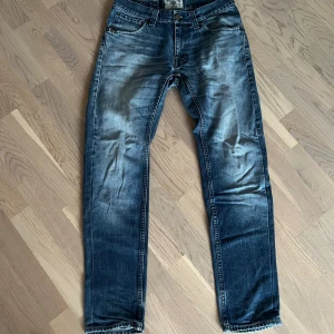 Blå jeans från Tiger Of Sweden med raka ben - Säljer ett par blå jeans från Tiger Of Sweden med snyggt slitna detaljer och raka ben. Jeansen har klassisk femficksdesign, normal midja och tydliga kontrastsömmar. Materialet är robust denim i bomull och passformen är normal. Perfekta för dig som gillar en avslappnad stil. OBS! (Inner längd 76cm)