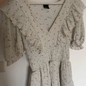 Beige blommig omlottklänning S - Supersöt beige kortklänning från Gina Tricot med små blommor i grönt och vitt. Klänningen har v-ringning, volangdetaljer och puffiga korta ärmar. Smockad midja för en snygg passform och omlottkänsla framtill. Perfekt för sommarens alla tillfällen!