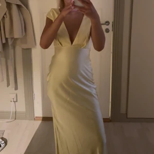 Ljusgul långklänning med djup v-ringning - Elegant långklänning i ljusgul satinlook med djup v-ringning och korta ärmar. Klänningen har en figurnära passform och släpper snyggt längs kroppen. Perfekt för dig som vill sticka ut med en stilren och modern look.