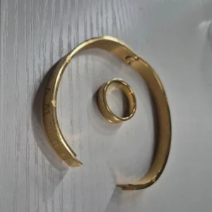 Guldigt armband och ring med romerska siffror - Snyggt set med ett guldigt armband och matchande ring, båda med ingraverade romerska siffror runt om. Armbandet är stelt och har en stilren design, medan ringen är bred och har samma mönster. Perfekt för dig som gillar statement-smycken med unik detalj.