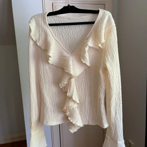 Beige blus med volang och v-ringning - Säljer en beige blus med lång ärm och v-ringning. Blusen har en snygg volangdetalj längs halsringningen och framtill, samt ett krinklat tyg som ger en cool struktur. Perfekt för dig som gillar romantiska och trendiga detaljer.