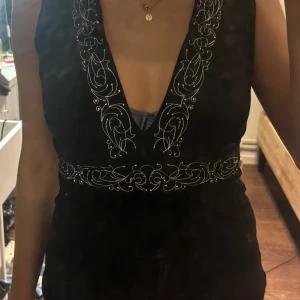 Svart ärmlös topp med broderi - Svart ärmlös topp med djup v-ringning och snygga vita broderade detaljer längs ringningen och midjan. Toppen har en figurnära passform och är tillverkad i ett mjukt material som känns skönt mot huden. Perfekt för dig som vill sticka ut med coola detaljer.