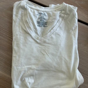 Vit v-ringad t-shirt från Polo Ralph Lauren - Vit t-shirt från Polo Ralph Lauren i klassisk passform, tillverkad i mjuk bomull. T-shirten har v-ringad hals och den ikoniska svarta Polo-loggan broderad på bröstet. Perfekt för en clean och stilren look.