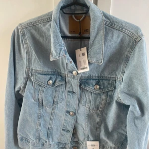 Ljusblå jeansjacka från Lager 157 - Snygg ljusblå jeansjacka från Lager 157 med klassisk krage, bröstfickor med knappar och raka sömmar. Jackan har långa ärmar och silverfärgade knappar framtill. Perfekt för dig som vill ha en tidlös och cool look.
