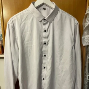 Vit klassisk skjorta med svarta knappar - Säljer en stilren vit skjorta med svarta knappar och klassisk krage. Skjortan har lång ärm och är tillverkad i ett slätt bomullstyg. Perfekt för dig som gillar en clean och enkel look.