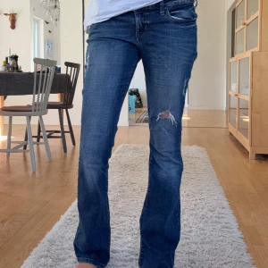 Blå bootcut jeans från Guess - Säljer ett par blå jeans från Guess med bootcut-snitt och klassisk femficksdesign. Jeansen har slitningar och hål på ena benet för en cool, avslappnad look. Materialet är jeans i bomull och de har en snygg mellanblå tvätt med detaljerade sömmar och Guess-logga på bakfickan.