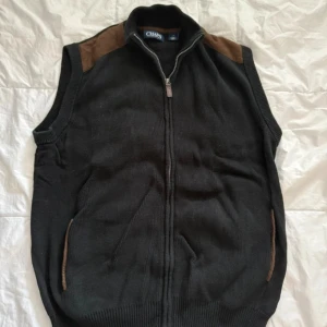 Svart stickad väst från Chaps - Svart stickad väst från Chaps med bruna mockadetaljer på axlarna och vid fickorna. Västen har hel dragkedja framtill och ribbade kanter. Var tyvärr lite för stor för mig därav säljes.