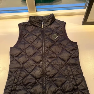 Svart quiltad dunväst från Gant - Quiltad svart dunväst från Gant med hög krage och dragkedja framtill. Västen har två fickor med dragkedja och en snygg Gant-logga på bröstet. Perfekt för lager-på-lager och stilren look.