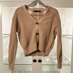 Beige stickad kofta från 157 XS - Snygg beige stickad kofta från 157 i storlek XS. Koftan har ribbad struktur, v-ringning och tre mörka knappar framtill. Långärmad och tillverkad i mjuk bomull, perfekt för lager-på-lager. Enkel och clean design som funkar till många olika stilar.