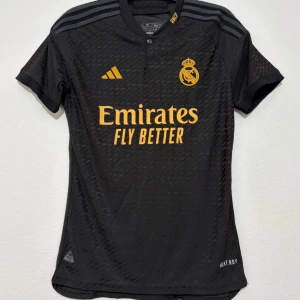 Real Madrid svart fotbollströja Adidas M - Snygg svart Real Madrid fotbollströja från Adidas med gula detaljer och klubbmärke på bröstet. Tröjan har korta ärmar, rund hals och diskret mönster i tyget. Tillverkad i lätt och ventilerande material, perfekt för träning eller match.