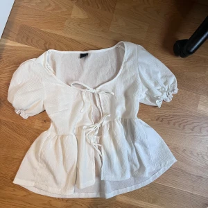 Vit blus med knyt från Gina Tricot - Supersöt vit blus från Gina Tricot med puffärmar och två knyt framtill. Blusen har en lätt rynkad struktur, markerad midja och rundad halsringning. Perfekt för dig som gillar romantisk och trendig stil.
