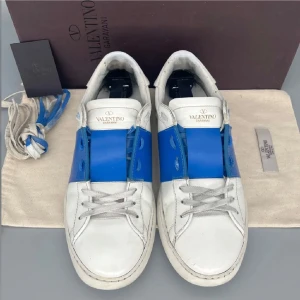 Valentino Garavani Open sneakers blå/vit - Snygga Valentino Garavani Open sneakers i vitt skinn med en bred blå rem över mitten. Klassisk låg modell med rund tå, platt sula och grå snörning. Kommer med originalkartong, dustbag och extra skosnören. Perfekta för dig som gillar exklusiva och trendiga sneakers.