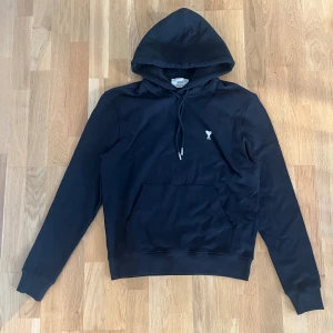 Svart hoodie från Ami Paris - Svart hoodie från Ami Paris med klassisk huva och snörning. Tröjan har en liten broderad logga på bröstet och en stor magficka framtill. Tillverkad i mjuk bomull, perfekt för en chill och stilren look.