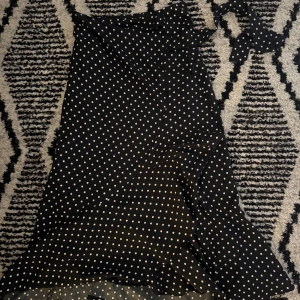 Svart prickig omlottkjol Gina Tricot - Supersöt svart omlottkjol från Gina Tricot med vita prickar och volangkant. Kjolen har knytband i midjan och är i en lätt och luftig polyester. Perfekt för dig som gillar en lekfull och trendig stil.