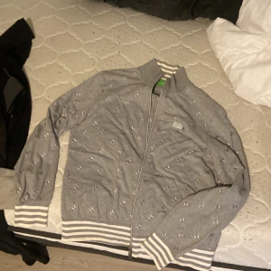 Grå ziptröja från BOSS med mönster - Säljer en grå långärmad ziptröja från BOSS med diskret mönster över hela tröjan. Tröjan har vita och grå ränder på muddar och krage, samt dragkedja framtill. Perfekt för dig som gillar stilren och sportig look.