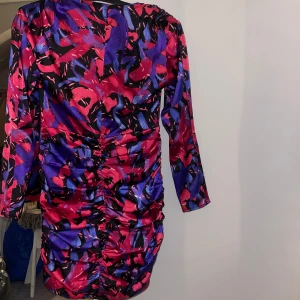 Färgstark kort klänning från Gina Tricot - Säljer en kort klänning från Gina Tricot med ett färgstarkt abstrakt mönster i rött, lila och svart. Klänningen har långa ärmar, v-ringning och snygg rynkdetalj framtill. 