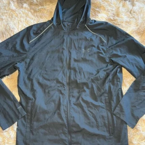 Svart vindjacka från Under Armour - Svart vindjacka från Under Armour med huva och hel dragkedja. Jackan har reflekterande detaljer på axlarna, meshfoder och logga på bröstet samt stort UNDER ARMOUR-tryck på ryggen. Perfekt för träning utomhus. Materialet är lätt och vattenavvisande.