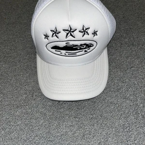 Vit truckerkeps från Corteiz med stjärnor - Vit truckerkeps från Corteiz med svart broderad grafik och stjärnor framtill, samt broderade stjärnor på sidan. Kepsen har mesh-bakstycke för extra ventilation och justerbar passform. Perfekt accessoar för en streetwear-look.