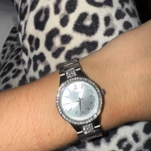 Silverfärgad klocka från Regal med stenar - Snygg klocka från Regal i silverfärg med glittrande stenar runt urtavlan och på armbandet. Klockan har en rund urtavla i silver och ett klassiskt metallarmband. Perfekt accessoar för att lyfta din stil.