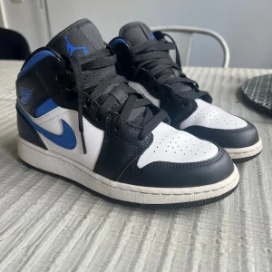 Nike Air Jordan 1 Mid svart/blå - Säljer ett par Nike Air Jordan 1 Mid sneakers i svart, vit och blå. Skorna har klassisk snörning, rund tå och platt sula. 