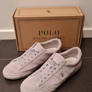 Grå mocka sneakers från Polo Ralph Lauren - Snygga grå sneakers från Polo Ralph Lauren i mjuk mocka med klassisk snörning och rund tå. Diskret Polo-logga på sidan och polo märke bak på sulan. Insidan har också ett Polo-tryck på innersulan. Dessa skor passar perfekt för dig som klär dig med stil, med den fina gråa färgen kan man styla skorna med flera olika outfits. Skorna har bara använts ett fåtal gånger på sommaren. 