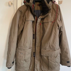 Beige parkajacka från Dressmann M - Parkajacka i beige från Dressmann med huva och stora fickor framtill. Jackan har röd snörning vid huvan, dragkedja och knappar, samt justerbara muddar vid ärmslut. På ryggen finns tre svarta punkter som knappt syns,men ni ser på sista bild .
