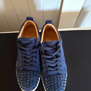 Blå sneakers Christian Louboutin - Säljer ett par blå sneakers från Christian Louboutin med coola nitar på tån och snörning. Skorna har en rund tå och vit platt sula. Insidan är beige och materialet känns lyxigt, troligen skinn eller mocka. Perfekt för dig som vill sticka ut med exklusiva detaljer.