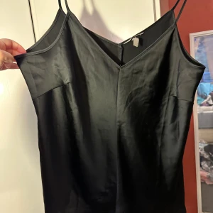 Svart satin linne från H&M strl: XL - Svart linne från H&M i storlek XL med smala axelband och v-ringad hals. Materialet är glansigt satin som ger en lyxig känsla. Linnet  är kort och har en enkel, stilren design som passar perfekt för dig som gillar minimalistisk stil. Obs, finns defekter ev dragning i tyget samt ena axelbandet har gått upp bäver sys. Annars i fint skick förutom de.