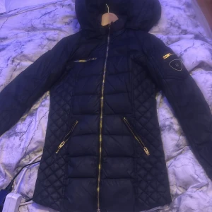 Rockandbluejacka  - Svart dunjacka från Rockandblue med huva och snygga guldfärgade dragkedjor. Jackan har quiltade partier på sidorna och ärmarna, samt flera fickor med dragkedja. Har ett litet hål där bak som går att sy 