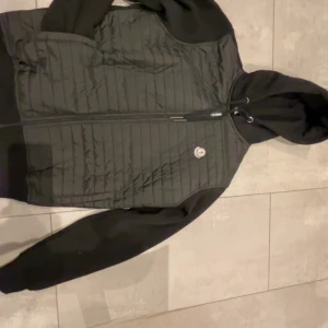 Svart Moncler pufferjacka med huva - Säljer en svart pufferjacka från Moncler i storlek M. Jackan har quiltad framsida, mjuka ärmar och huva med snörning. Dragkedja framtill och Moncler-logga på bröstet. Perfekt för kyliga dagar och riktigt snygg streetstil.